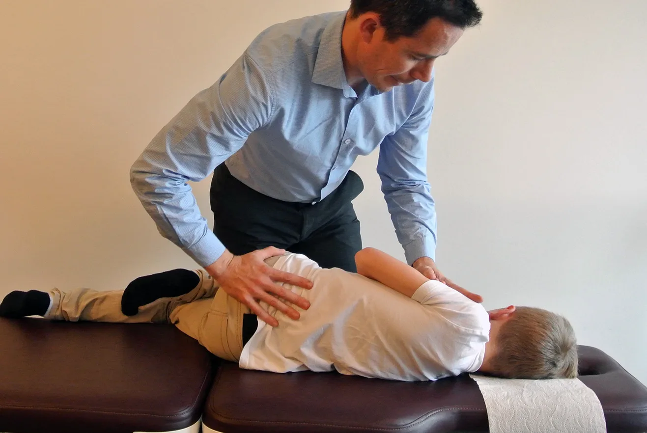 A chiropractor in a blue shirt performing a gentle hands-on adjustment on a child on the treatment table